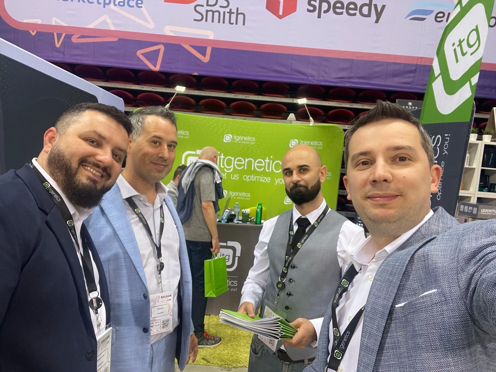 A selfie of four people wearing IT Genetics lanyards in front of the green stand of IT Genetics, Planograma's parent company, one holding a stack of green brochures