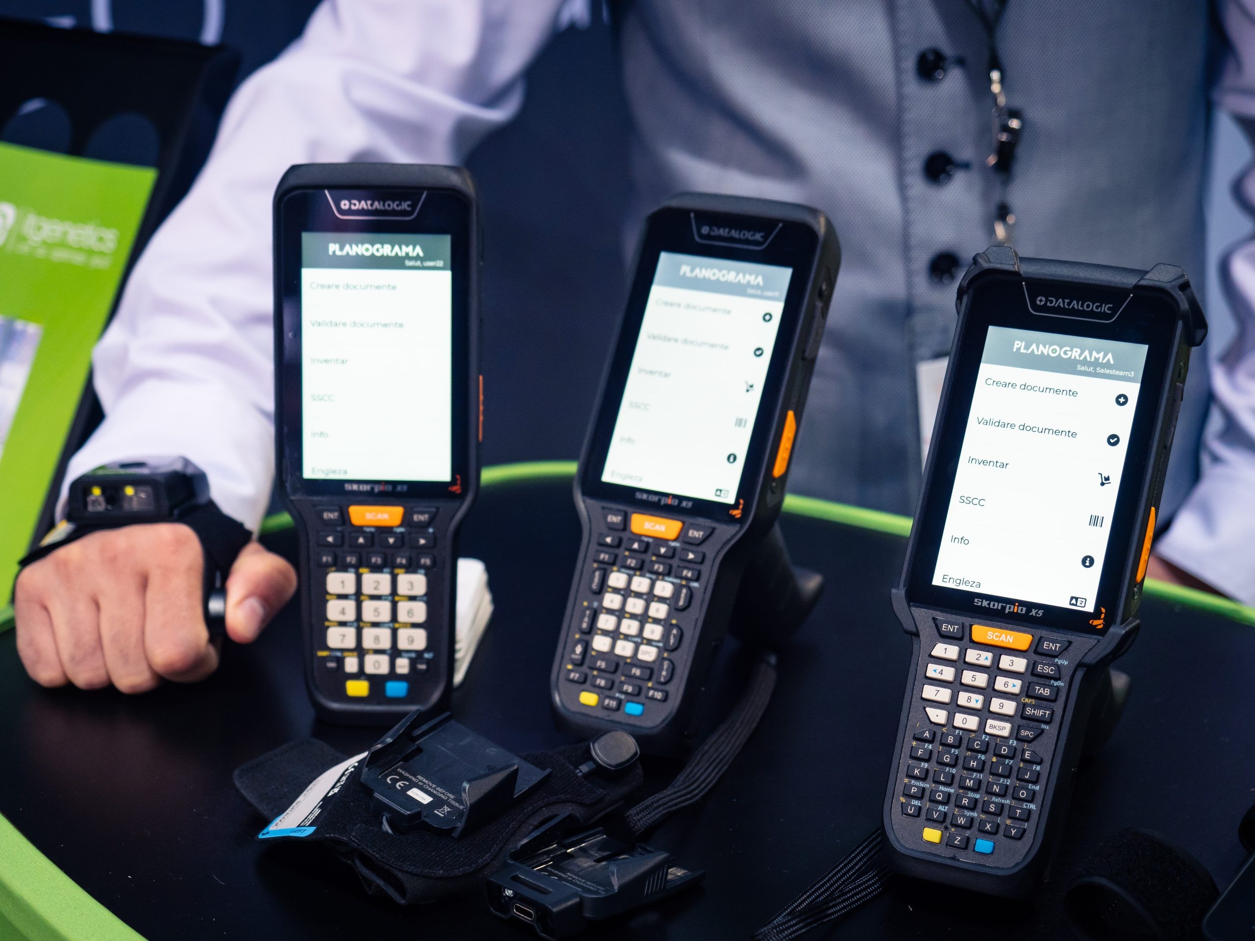 Three Datalogic handheld computers running the Planograma app on the stand's counter, beside a wearable barcode scanner on a staff member's hand