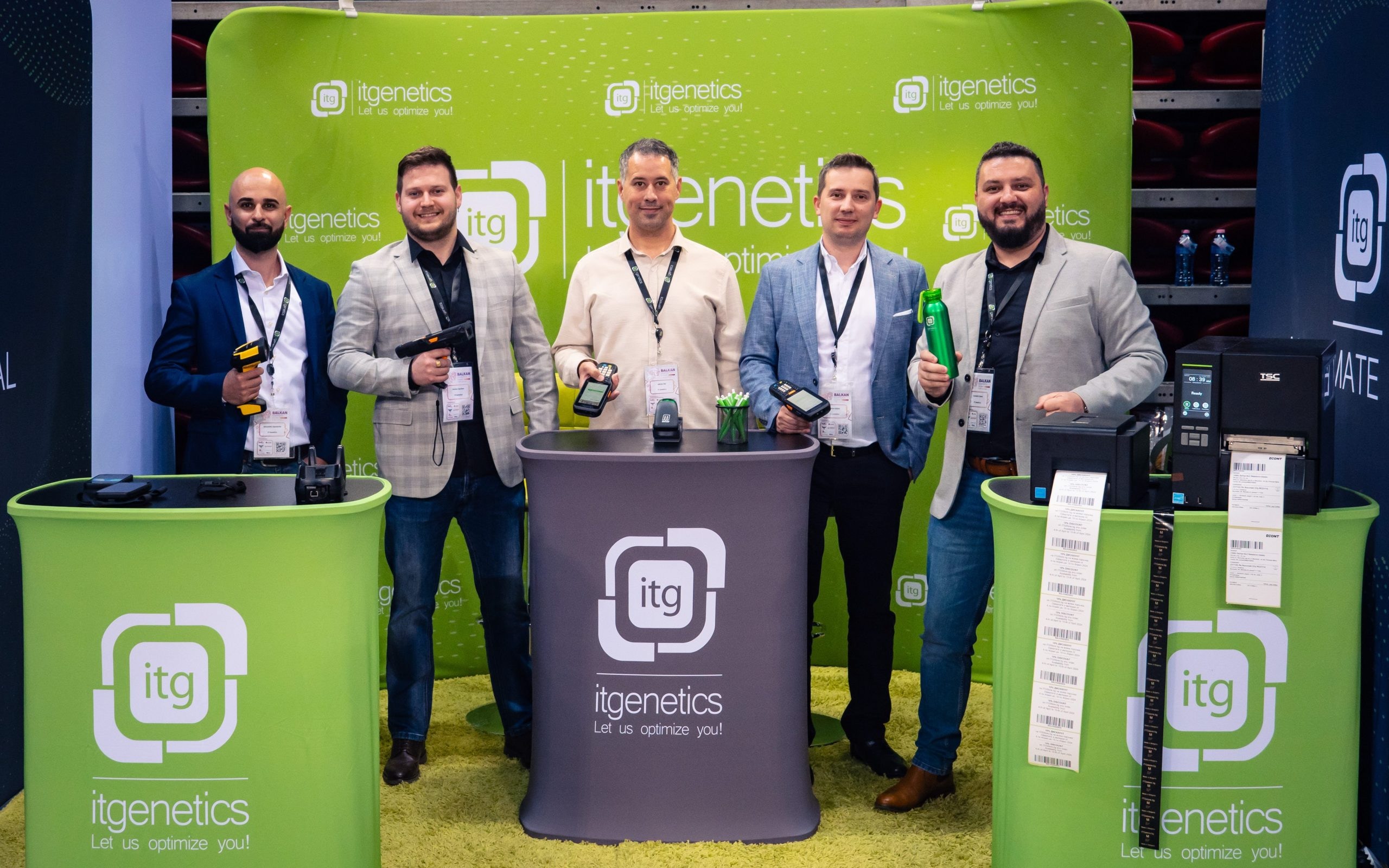 Five people posing at the IT Genetics stand with handheld barcode scanners and mobile computers, next to label printers and strips of printed barcode labels